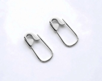 Könnte beinhalten: Ein Paar silberfarbene Ohrringe in Form von Sicherheitsnadeln mit polierter metallischer Oberfläche. Jeder Ohrring besteht aus einer U-förmigen Drahtschlaufe und einem sicheren Verschlussmechanismus an der Oberseite mit glatter Optik.