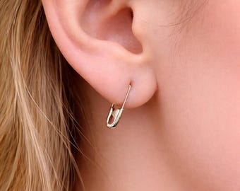 Könnte beinhalten: Ein goldfarbener Sicherheitsnadel-Ohrring, der im Ohrläppchen getragen wird. Der Ohrring hat die Form einer klassischen Sicherheitsnadel mit polierter Metalloberfläche, einem funktionalen Verschlussmechanismus unten und einem dünnen Drahtstift.