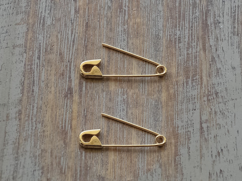May include: Two gold-toned metal safety pins are positioned horizontally, one above the other. Each pin features a smooth, polished finish with a classic coiled spring mechanism at one end and a secure clasp closure at the other end.