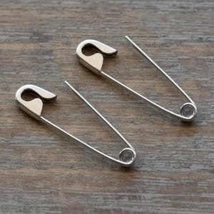May include: Two silver-toned metal safety pins with a polished finish. Each pin features a classic U-shaped wire design with a secure clasp mechanism at one end and a coiled spring loop at the other, presented in an open and functional position.