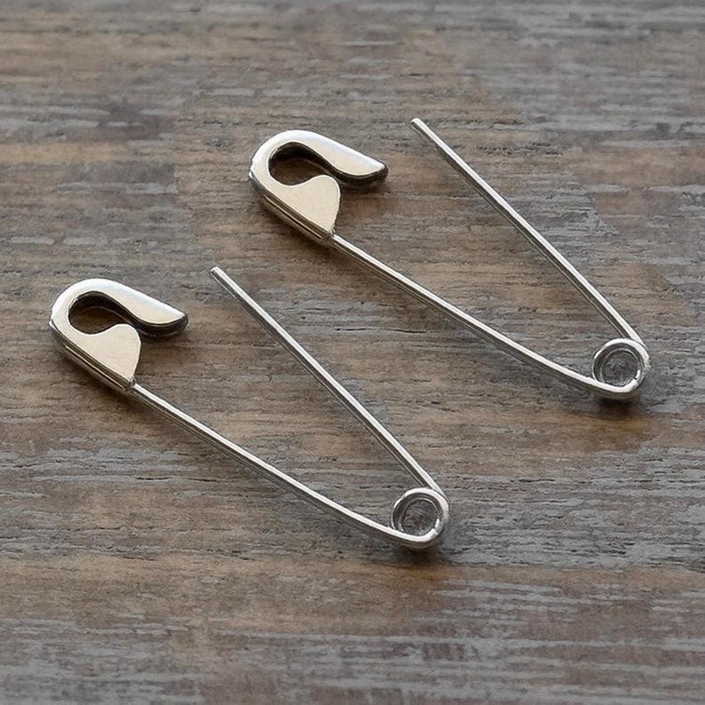 May include: Two silver-toned metal safety pins with a polished finish. Each pin features a classic U-shaped wire design with a secure clasp mechanism at one end and a coiled spring loop at the other, presented in an open and functional position.