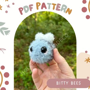 May include: A close-up of a hand holding a small, gray, fuzzy toy bee with black eyes. The toy is being held against a green, leafy background. The text "PDF PATTERN" is in the upper left corner of the image, and the text "BITTY BEES" is in the lower right corner of the image.