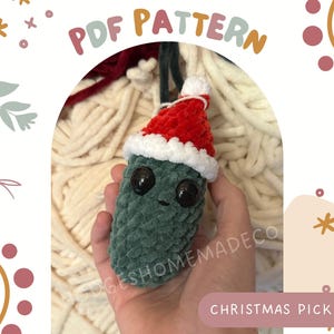 Pode incluir: Um enfeite de Natal de picles verde em crochê com um chapéu de Papai Noel vermelho e branco em crochê. O enfeite tem olhos pretos e uma boca sorridente. O enfeite está sendo segurado na mão de uma pessoa.