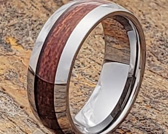May include: A polished silver ring with a central band of reddish-brown wood. The ring has a smooth, reflective surface, and the wood grain is visible. The ring is likely a wedding band or fashion accessory.