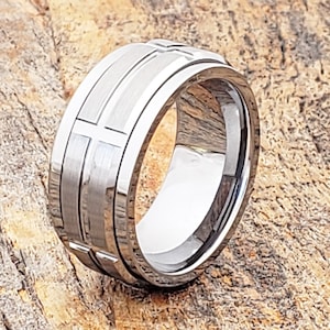Puede incluir: Un anillo plateado con un acabado de metal cepillado. El anillo presenta una serie de diseños rectangulares y en forma de cruz alrededor de su circunferencia. El anillo se muestra sobre una superficie de madera.