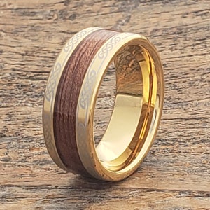 Op de afbeelding: Een goudkleurige ring met een middenband in houtnerf en Keltische knoopontwerpen op de randen. De ring heeft een gepolijst, reflecterend oppervlak en het hout lijkt donkerbruin van kleur.