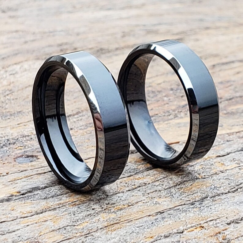 May include: Two black and silver wedding bands with beveled edges. The bands are made of tungsten carbide and have a polished finish.
