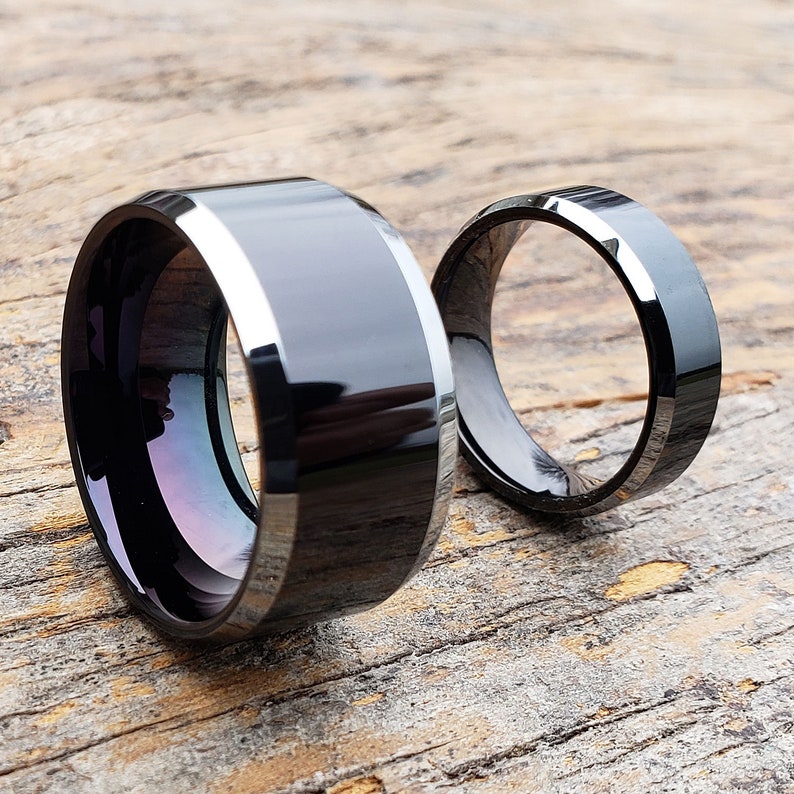 May include: Two black and silver wedding bands. The band on the left is wider and has a beveled edge. The band on the right is thinner and has a smooth finish.