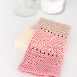 May include: A knitted washcloth in shades of pink and beige, with a rectangular bar of soap. The washcloth has a textured, handmade appearance, with a decorative openwork design near the top. The items are arranged on a white surface.