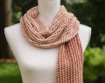 May include: A peach-colored, open-weave scarf is draped on a mannequin. The scarf has a textured, knitted appearance with a rectangular shape. The scarf is displayed against a blurred green background, suggesting an outdoor setting. The scarf's color is a blend of light peach and darker peach.
