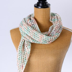 Könnte beinhalten: Ein gehäkelter Schal in Grün-, Rosa- und Weißtönen mit einem zarten Lochmuster. Der Schal ist um eine mit Sackleinen bezogene Schaufensterpuppe gelegt.