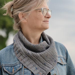 May include: A woman wearing a denim jacket and a gray knitted scarf with blue and red flecks. She is looking off to the side and has gray hair.