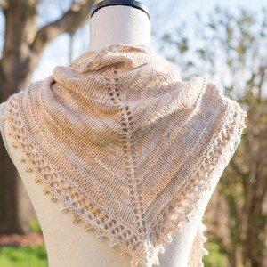 May include: A beige knitted shawl with a delicate lace pattern, draped over a white mannequin. The shawl is displayed against a blurred background of green grass and trees.