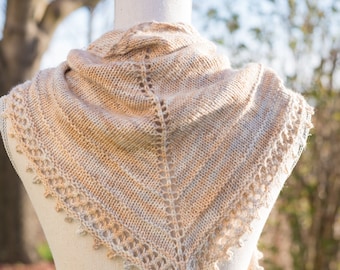 May include: A beige knitted shawl with a delicate lace pattern, draped over a white mannequin. The shawl is displayed against a blurred background of green grass and trees.