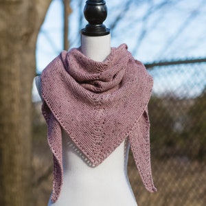 Puede incluir: Un chal de punto rosa polvoriento con un delicado patrón de encaje drapeado sobre un maniquí blanco.