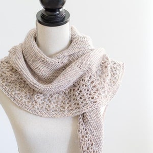 May include: A beige knitted shawl with a delicate lace pattern draped over a white mannequin.