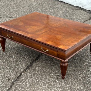 May include: Rectangular wooden coffee table with a rich, warm brown finish. The table features a patterned top, two drawers with brass-toned handles, and decorative, fluted legs. A classic piece of furniture.