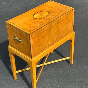 May include: A light-colored wooden chest with a decorative inlaid oval on the lid, set atop a matching stand. The chest features a brass handle and is constructed with a light wood grain. The stand has four legs with a cross-brace for support.