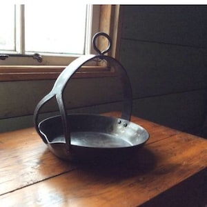 May include: A black metal pan with a handle that forms a loop. The pan is sitting on a wooden table.