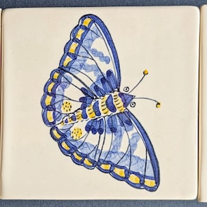 May include: Three square ceramic tiles, each featuring a hand-painted butterfly illustration. The left tile shows a butterfly with orange and grey wings. The centre tile displays a blue and yellow butterfly. The right tile depicts a yellow and blue butterfly.