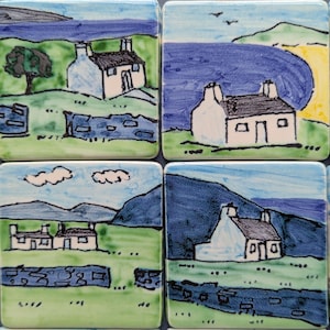 May include: A set of four square ceramic tiles featuring hand-painted illustrations of white cottages in coastal landscapes. Each tile depicts green fields, blue water, stone walls, and small houses with dark roofs under blue skies and clouds.