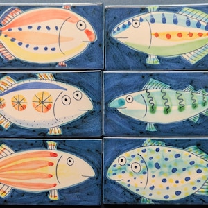 May include: Six hand-painted ceramic tiles with quirky fish designs. Each fish has a unique pattern in shades of orange, blue, yellow, and green. The tiles are set against a dark blue background, creating a striking contrast. Ideal for kitchen or bathroom decoration.