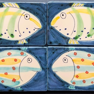 May include: Four ceramic tiles with hand-painted fish designs. Two tiles feature fish with yellow and green stripes, while the other two have blue and yellow stripes with red spots. The background is a dark blue, textured pattern.