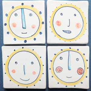 May include: Four square ceramic coasters with whimsical sun-like faces. Each coaster features a white background, a yellow sun outline, blue accents, and a unique facial expression with pink cheeks.
