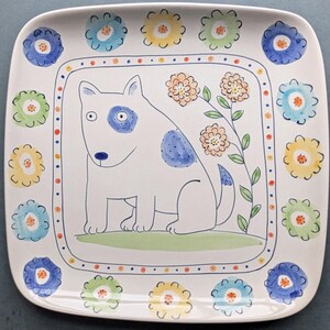 May include: A white ceramic square serving platter with a hand-painted design of a dog with blue spots and a green plant with orange flowers. The border of the platter is decorated with a row of colorful flowers.
