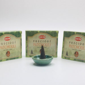 Op de afbeelding: Drie dozen HEM Precious Patchouli wierookkegels, met een groene achtergrond en gouden letters. De dozen zijn in een rij geplaatst, met een groene keramische wierookbrander in het midden.