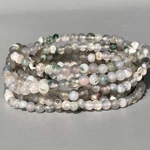 Könnte beinhalten: Ein Stapel Armbänder aus Perlen. Die Armbänder bestehen aus kleinen, runden, halbtransparenten Perlen in Grau-, Weiß- und Blassrosatönen. Einige Perlen haben grüne Einschlüsse. Die Armbänder sind auf einer grauen Oberfläche gestapelt.