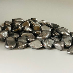 Può includere: Un mucchio di pietre di ematite lisce e di colore grigio scuro, levigate a forma di ciottoli. Le pietre sono di diverse dimensioni e forme e hanno un aspetto metallico.
