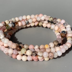 May include: A collection of delicate beaded bracelets. The bracelets are made of small, round beads in shades of pink, cream, and brown. The beads are strung together to form multiple strands.