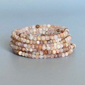 May include: A stack of beaded bracelets in shades of pink, cream, and brown. The small, round beads are strung together, creating a flexible and layered accessory. The bracelets are displayed against a gray background.