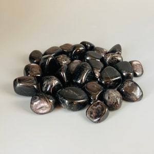 May include: A pile of black obsidian tumbled stones with a slight iridescent sheen. The stones are smooth and polished, and they are arranged in a random pattern.