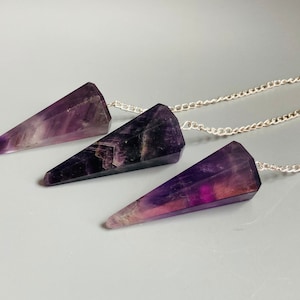 Könnte beinhalten: Drei Amethyst-Kristallpendel mit Silberketten. Jedes Pendel hat eine dreieckige Prismenform mit verschiedenen Lila- und Weißtönen. Die Kristalle sind an filigranen Silberketten aufgehängt.
