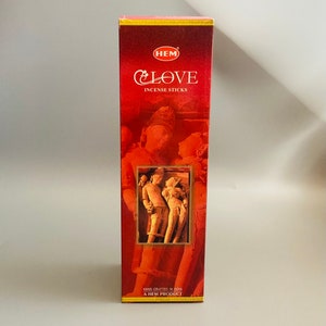 Puede incluir: Una caja roja de inciensos HEM CLOVE. La caja tiene un borde dorado y una imagen de una pareja en un abrazo amoroso. El texto "HEM CLOVE INCENSE STICKS" está impreso en la caja. La caja también indica "HAND CRAFTED IN INDIA A HEM PRODUCT."