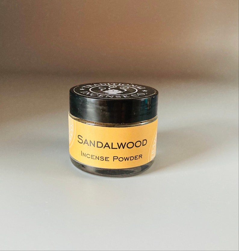 May include: A small glass jar with a black lid and a yellow label that reads "Sandalwood Incense Powder". The jar is filled with a dark brown powder.