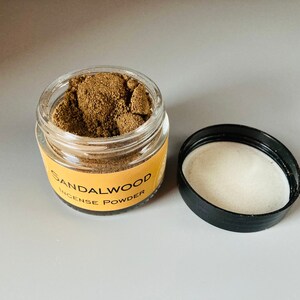 May include: A small glass jar with a black lid filled with brown sandalwood incense powder. The jar has a yellow label that reads "Sandalwood Incense Powder".
