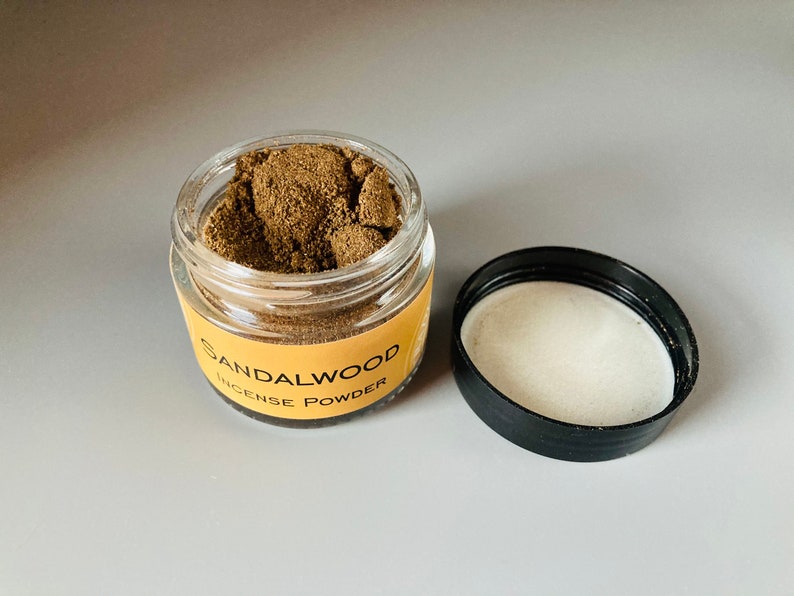 May include: A small glass jar with a black lid filled with brown sandalwood incense powder. The jar has a yellow label that reads "Sandalwood Incense Powder".