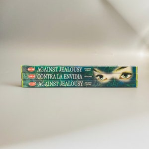 Puede incluir: Tres cajas de inciensos en forma de varillas apiladas una encima de la otra. Las cajas son verdes y azules con texto blanco. La caja superior dice "Against Jealousy" y la caja inferior dice "Contra La Envidia".