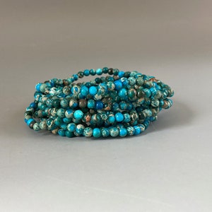 Puede incluir: Una colección de pulseras de cuentas turquesas y verde azulado. Las pulseras están hechas de pequeñas cuentas redondas con una mezcla de colores, incluyendo azul, verde y marrón. Las pulseras están apiladas.