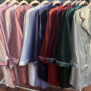 Puede incluir: Una colecci&oacute;n de diez camisas de dormir de sat&eacute;n en varios colores, incluyendo beige, rosa, azul claro, azul marino, burdeos, verde y verde claro. Las camisas est&aacute;n colgadas en perchas y tienen un frente abotonado y mangas largas.