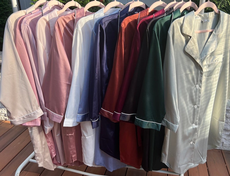 Puede incluir: Una colecci&oacute;n de diez camisas de dormir de sat&eacute;n en varios colores, incluyendo beige, rosa, azul claro, azul marino, burdeos, verde y verde claro. Las camisas est&aacute;n colgadas en perchas y tienen un frente abotonado y mangas largas.
