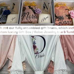 Peut inclure: Trois bo&icirc;tes cadeaux contenant des robes en satin. Les bo&icirc;tes sont blanches avec une bordure dor&eacute;e et les robes sont rose, blanche et bleu clair. Chaque robe pr&eacute;sente une grande lettre avec des motifs floraux et les mots "Bride" ou "Bridesmaid". Les bo&icirc;tes comprennent &eacute;galement un petit coffret &agrave; bijoux et un gobelet.
