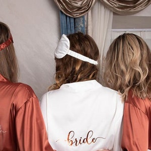 Puede incluir: Tres mujeres con batas de sat&eacute;n a juego con las palabras "bride" y "bridesmaid" bordadas en la espalda. La bata de la novia es blanca y las batas de las damas de honor son de color &oacute;xido.