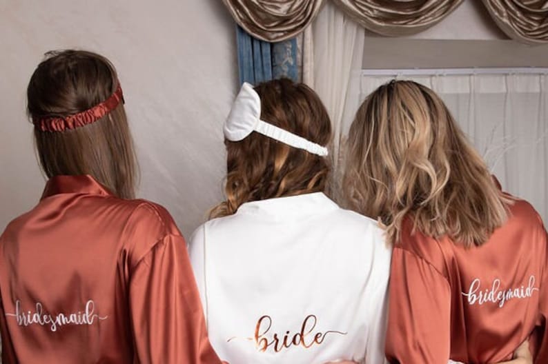 Puede incluir: Tres mujeres con batas de sat&eacute;n a juego con las palabras "bride" y "bridesmaid" bordadas en la espalda. La bata de la novia es blanca y las batas de las damas de honor son de color &oacute;xido.