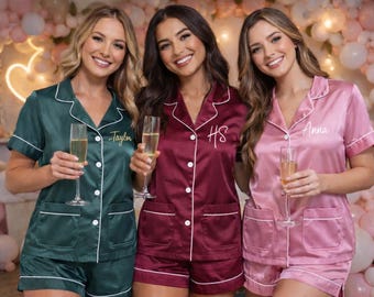 May include: Three women wearing matching satin pajama sets in emerald green, burgundy, and pink, each holding a champagne flute. The pajamas have white piping and personalized names or initials embroidered on the chest pocket.
