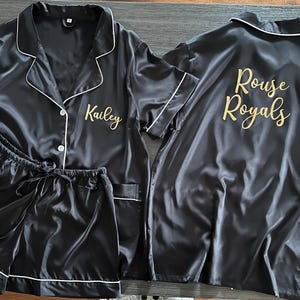 May include: Black satin pajama set with white trim, including a button-down shirt and shorts. The shirt is embroidered with "Kailey" in gold, and the back of another shirt reads "Rouse Royals".
