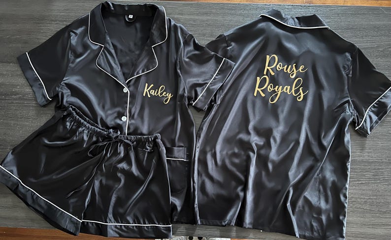May include: Black satin pajama set with white trim, including a button-down shirt and shorts. The shirt is embroidered with "Kailey" in gold, and the back of another shirt reads "Rouse Royals".